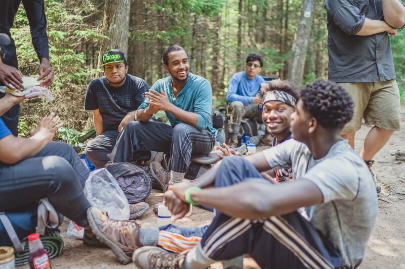 A group of young men are gathered in a wooded area, possibly on a hike or camping trip. They are sitting and standing around, engaged in conversation and appearing relaxed. The atmosphere seems casual and friendly, suggesting they are enjoying their time together in nature. The scene captures a moment of camaraderie and connection among the group.